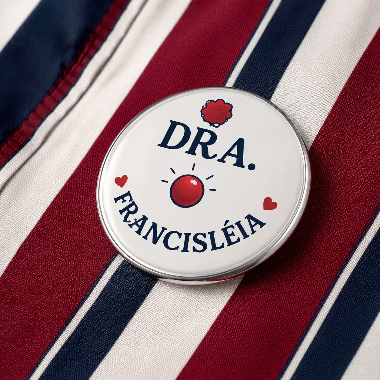 Broche 'Dra. Francisléia' sobre tecido listrado em vinho, branco e marinho, ao lado de um nariz de palhaço vermelho