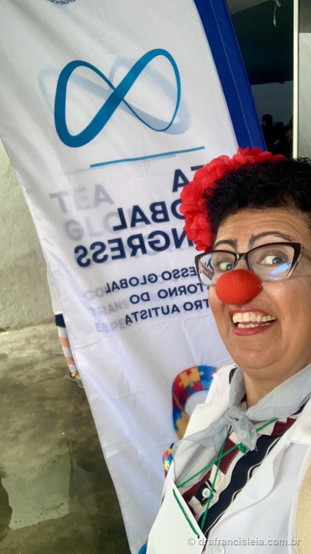 Dra. Francisléia sorrindo em selfie ao lado do banner do Congresso Global do Transtorno do Espectro Autista