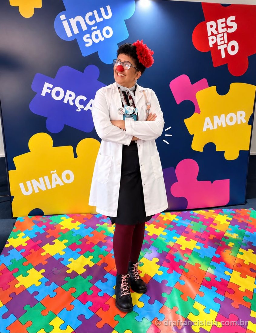 Dra. Francisléia de jaleco e nariz vermelho diante de um painel de peças de quebra-cabeça com as palavras Inclusão, Respeito, Força, Amor e União