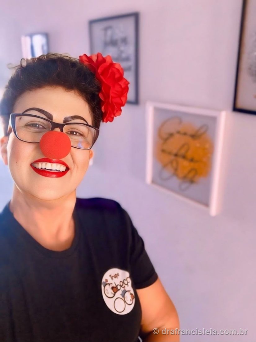 Selfie sorridente da Dra. Francisléia com nariz de palhaço vermelho, usando camiseta da Trupe Cabeça Oca